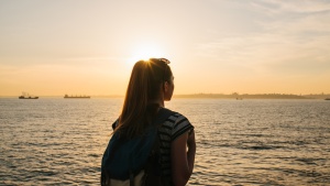 Une jeune touriste avec un sac à dos se tient près de la mer au coucher du soleil et regarde au loin.