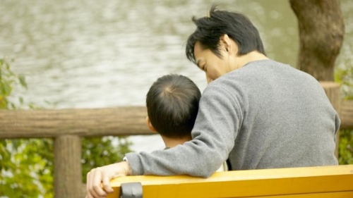 Un père et son fils s'assoient sur un banc au parc. Le père sourit en parlant à son fils.