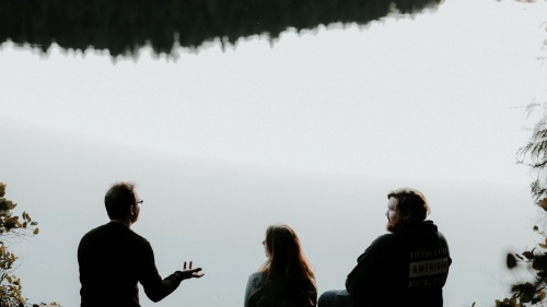Trois personnes sont assis au bord d'un lac — une personne parle aux autres qui l'écoutent