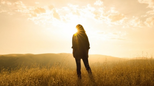 Une femme dans un champs qui regard le soleil au-dessous des montagnes