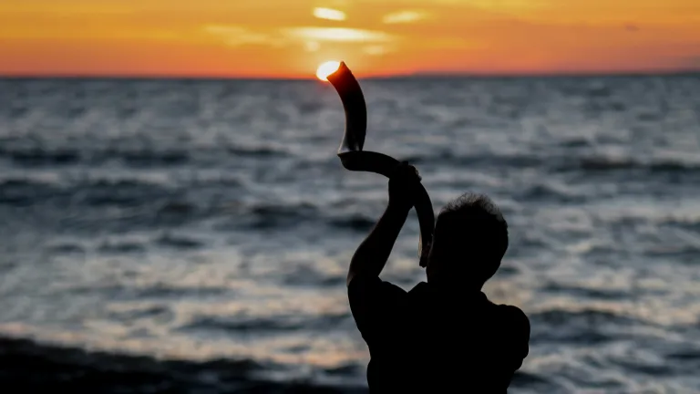 a silhouette of a person blowing into a shofar with sunset over the ocean in the background