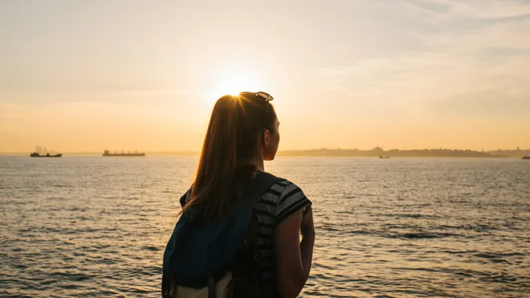 Une jeune touriste avec un sac à dos se tient près de la mer au coucher du soleil et regarde au loin.