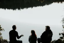 Trois personnes sont assis au bord d'un lac — une personne parle aux autres qui l'écoutent