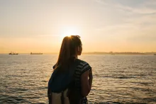 Une jeune touriste avec un sac à dos se tient près de la mer au coucher du soleil et regarde au loin.