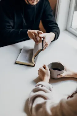 Une homme et une femme sont assis à une table et l'homme tourne la page d'une Bible.